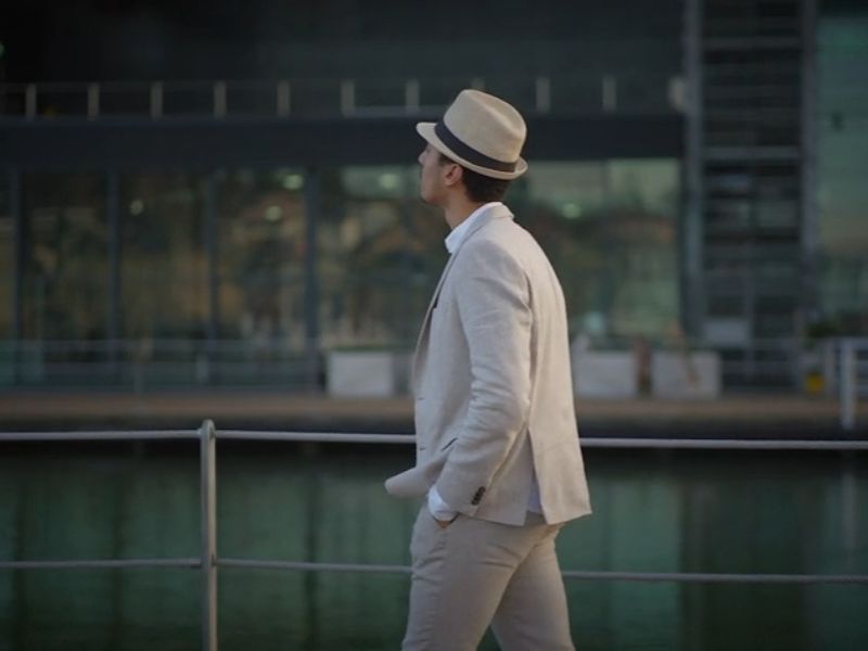 Muškarac u svetlom letnjem odelu i šeširu šeta pored vode.