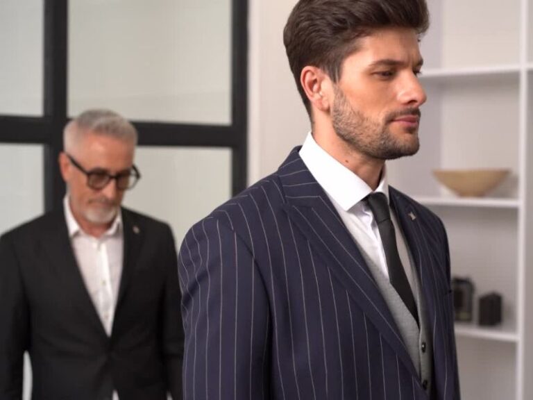 Na slici su dva muškarca obučena u elegantna muška odela. Muškarac u prvom planu nosi tamno plavo odelo sa belim prugama, belu košulju, sivi prsluk i crnu kravatu.