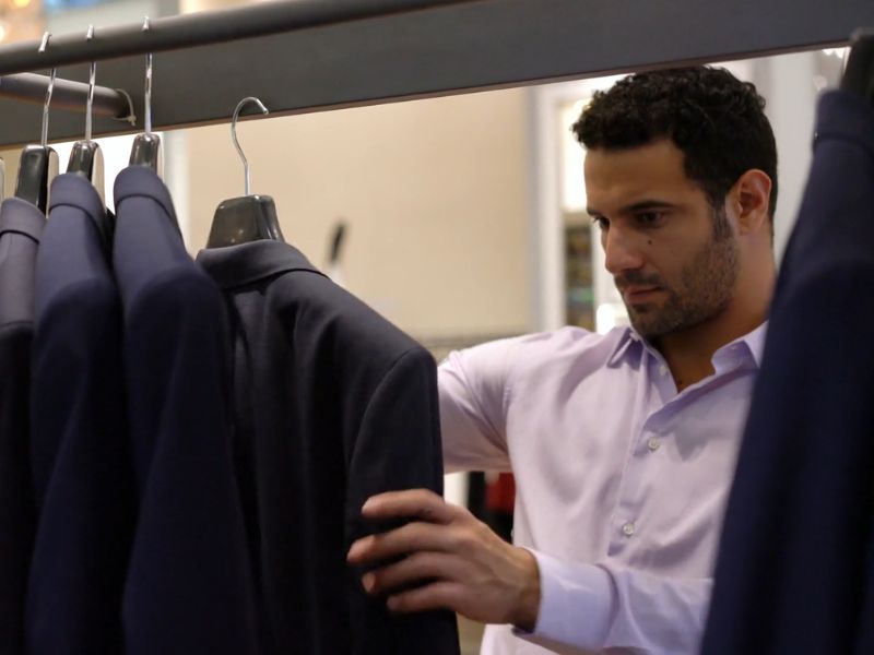 Muškarca koji bira muško odelo sa police u prodavnici. Okružen je tamnim odelima na ofingerima, dok pažljivo proučava jedan komad garderobe.