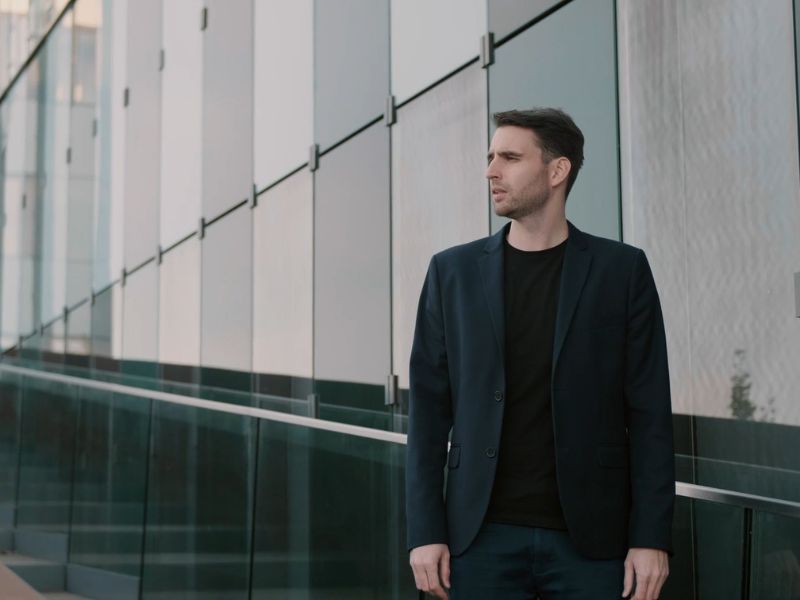Muškarac obučen u tamnoplavo casual odelo sa crnom majicom ispod, stoji pored moderne staklene zgrade, gledajući u daljinu.
